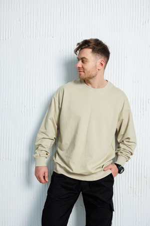 A man dressed in a comfortable beige sweatshirt along with stylish black pants is casually leaning against a clean white wall nearbyの写真素材