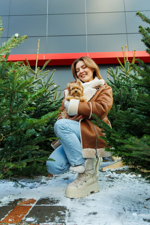 A cheerful woman in a stylish brown coat joyfully holds a small dog while sitting among beautifully decorated Christmas trees, capturing the warmth of winter festivitiesの写真素材