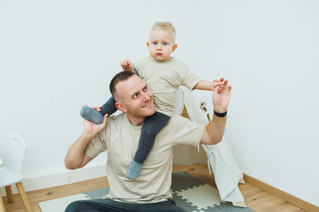 A joyful father shares a delightful moment with his beloved son, who gleefully rides on his shoulders in a bright and cheerful indoor setting filled with warmth and loveの写真素材