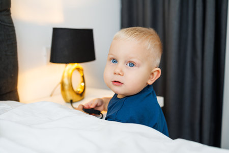 A charming toddler with bright blue eyes and short blonde hair is sitting on a cozy bed, looking with curiosity towards the camera in a beautifully designed modern bedroomの写真素材