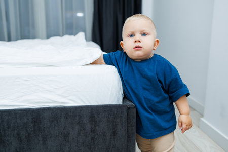 A charming little toddler stands next to a tidy bed in a stylish bedroom, showcasing an innocent curiosity that evokes feelings of warmth, pure joy, and heartwarming innocenceの写真素材