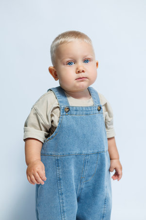 A cute little baby dressed in denim overalls and a light shirt stands confidently against a soft blue background, making it perfect for family or childrens projects and memoriesの写真素材
