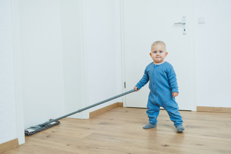 A cheerful toddler in a bright blue outfit pretends to use a vacuum cleaner, showing delight and eagerness to help with household chores and promote developmentの写真素材