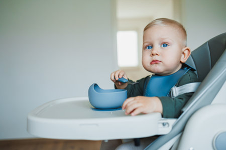 A cute baby with bright blue eyes sits happily in a high chair, eagerly eating from a blue bowl, with a minimalistic background creating a cozy mealtime atmosphereの写真素材