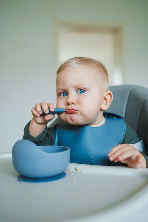 A charming baby with sparkling blue eyes and a messy mouth is joyfully feeding themselves with a spoon in a high chair, set against a bright kitchen backgroundの写真素材