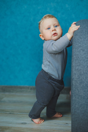 In a stylish space, a charming baby in cute clothes holds soft gray furniture against a vibrant blue wall, showcasing wonder and joy in a cozy area for family momentsの写真素材