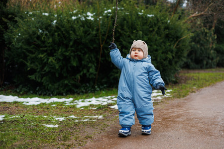 A cheerful toddler, dressed in a bright blue snowsuit and a warm hat, happily explores a snowy outdoor path, wielding a small stick during this lovely winter seasonの写真素材