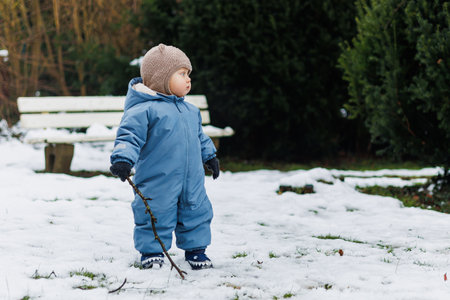 A joyful child in a blue winter suit explores the snowy outdoors, playfully holding a stick amidst lush greenery and cozy benches in a magical winter wonderlandの写真素材