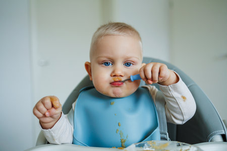 A cheerful baby sitting in a high chair, joyfully exploring food with a blue spoon. The bright setting enhances the charming moment of early selffeedingの写真素材