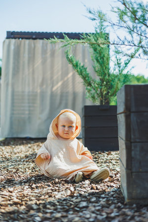A cute baby in warm clothes sits among colorful pebbles outdoors, framed by greenery, creating a charming scene of joy and innocent childhoodの写真素材