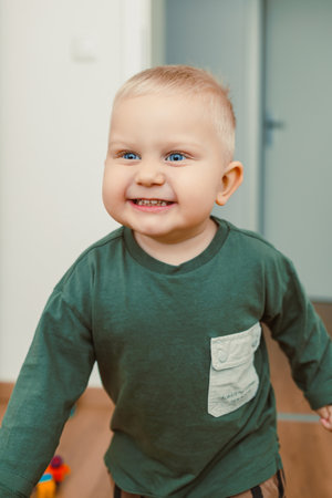A cheerful toddler with sparkling blue eyes and a big smile, in a vibrant green shirt, joyfully playing in a bright, modern living space filled with natural lightの写真素材