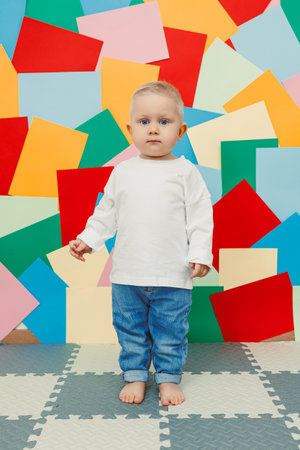 An adorable toddler in a white shirt and blue jeans stands barefoot against a vibrant geometric backdrop, perfect for capturing precious family and childrens momentsの写真素材