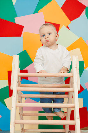 A lively toddler, full of curiosity, climbs a sturdy wooden ladder with a joyful smile, surrounded by vibrant, colorful geometric shapes in a playful atmosphereの写真素材
