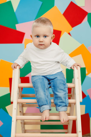 An adorable and charming baby joyfully sitting on a wooden ladder surrounded by vibrant colored shapes in the backdrop, capturing the essence of childhood exploration and pure joyの写真素材