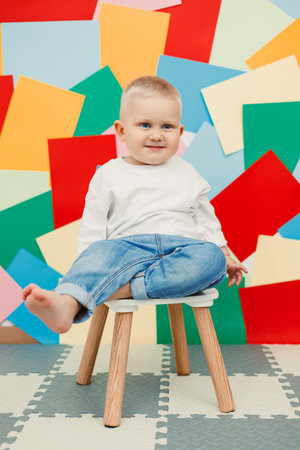 A cheerful toddler sits on a colorful stool, surrounded by playful paper shapes, embodying joyful childhood moments filled with laughter and vibrant imaginationの写真素材