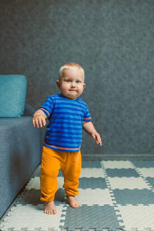 A cheerful toddler in a blue striped shirt and orange pants stands barefoot on a patterned floor, embodying innocent curiosity in a stylish, modern living spaceの写真素材