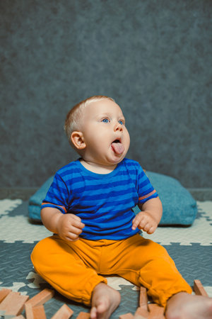 A cheerful toddler in bright blue and orange clothing sits on a soft mat, playfully sticking out their tongue, capturing a joyful, innocent moment filled with laughterの写真素材