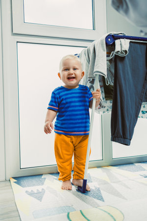 A joyful baby excitedly stands beside a colorful clothes rack in a beautifully sunlit room, perfectly capturing the pure innocence and delight of childhood in this adorable momentの写真素材