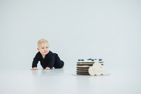 In a joyful and minimalistic setting, a delightful little baby wearing a black outfit crawls eagerly toward a delicious cake, radiating pure curiosity and sweetnessの写真素材