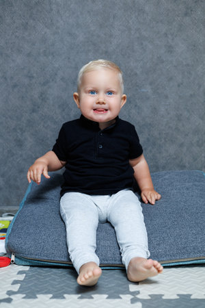 A joyful baby in a black polo and gray leggings sits on a fluffy cushion, showcasing a bright smile, perfect for capturing lasting memories with family and friendsの写真素材