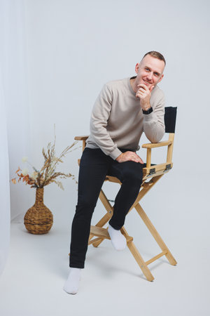 A cheerful young man confidently poses on a directors chair, showcasing his casual yet stylish outfit. This scene is perfect for lifestyle, fashion, or portrait photographyの写真素材
