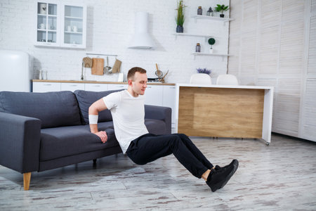 In a modern living room with stylish decor, a young man energetically does tricep dips on a sofa, showcasing home fitness options and promoting a healthy lifestyleの写真素材