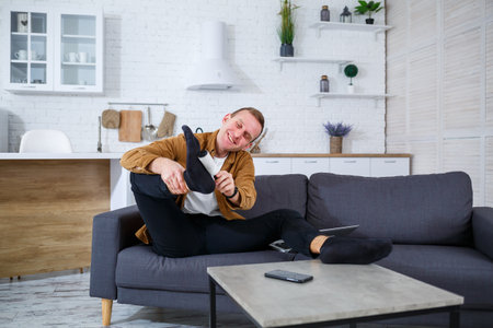 A young man relaxes on a stylish couch, adjusting his shoe for a perfect fit. The room's modern decor creates a warm atmosphere for unwinding after a long dayの写真素材