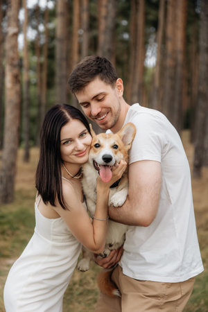 In a serene and beautiful forest, a joyful couple lovingly embraces their playful and lively corgi, capturing a truly heartfelt moment of love amidst the vibrant beauty of natureの写真素材