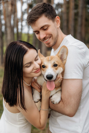 A joyful couple shares a beautiful and tender moment in a serene forest, accompanied by their charming corgi, radiating love and happiness amidst the warm sunlightの写真素材