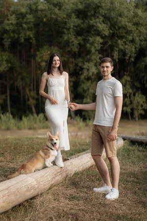 A couple stands together in a lush green setting, enjoying the day with their adorable corgi beside them. Perfect representation of love and companionship in nature.の写真素材