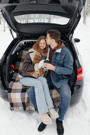In a picturesque winter wonderland, a couple happily shares warm drinks and sweet cuddles in their cars trunk, forging a truly cozy atmosphere that radiates warmth and deep loveの写真素材