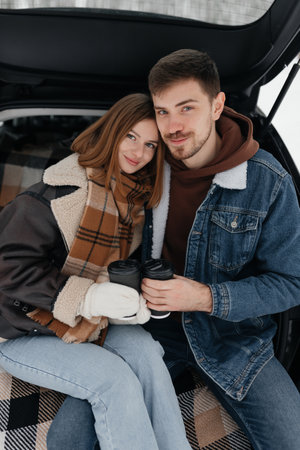 A stylish couple cozily snuggles together inside the back of a car, enjoying warm drinks as they embrace the stunning beauty of the winter scenery outside their windowの写真素材