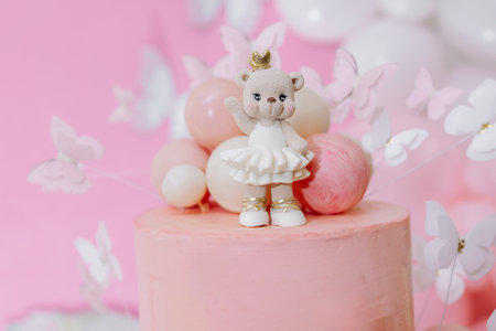 A pink birthday cake topped with a teddy bear figurine is surrounded by colorful balloons and butterflies, creating a joyful centerpiece for childrens celebrationsの写真素材
