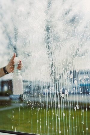 A hand grips a spray bottle to clean a window but leaves streaks. Behind the glass, the stunning beauty of nature is vividly visible, creating a beautiful contrastの写真素材