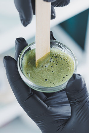 A detailed closeup shows a person mixing a vibrant green herbal mask in a glass bowl with a wooden stick, highlighting the art of natural skincare preparationの写真素材