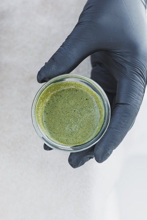 This image highlights a closeup of a vibrant green herbal mix in a clear glass jar, held by a gloved hand, symbolizing its use for natural skin care and wellnessの写真素材