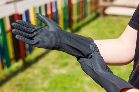 A persons hand is putting on black rubber gloves outdoors in a vibrant park with a colorful fence, illustrating cleanliness and protection from dirt and germsの写真素材