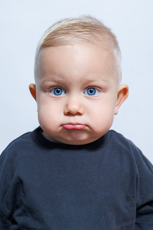A delightful and charming baby with a pouting expression that showcases mesmerizing striking blue eyes and wonderfully styled blonde hair, adorably dressed in a cozy black shirtの写真素材