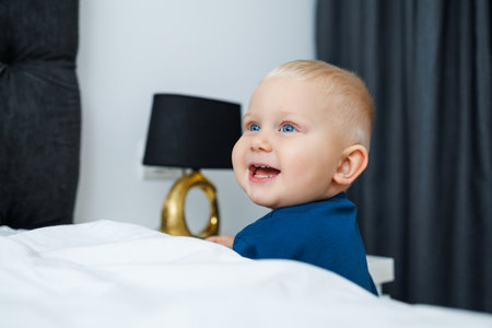 A cute baby with sparkling blue eyes and a joyful smile is sitting on a plush bed in a cozy bedroom, creating a heartwarming and serene atmosphere filled with loveの写真素材