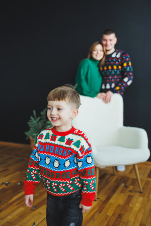 A cheerful and happy child, dressed in a colorful holiday sweater, stands joyfully in front of a loving family, fully embodying the festive spirit in a cozy home environmentの写真素材