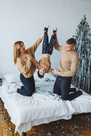 Joyful parents playfully hold their cheerful child upside down on a cozy bed, celebrating love and togetherness this festive holiday season with smiles and laughterの写真素材