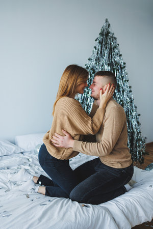 A young couple shares a tender moment on their bed, surrounded by a warm atmosphere and a beautifully decorated Christmas tree, capturing the essence of love and intimacy.の写真素材