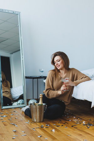 A joyful woman, radiating happiness, sits among colorful festive decorations, holding a sparkling champagne bottle in her stylish bedroom, embodying cozy celebrationsの写真素材