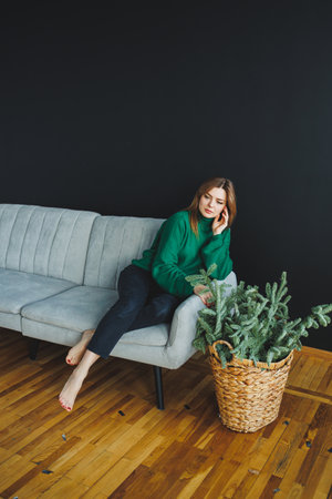 A fashionable young woman in a cozy green sweater sits on a chic grey couch, surrounded by vibrant green plants against an elegant black wall, creating a serene atmosphereの写真素材