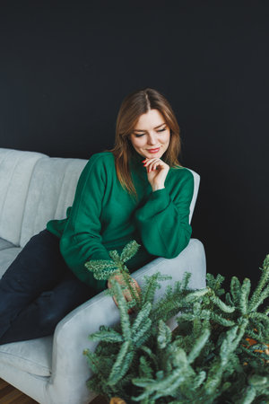 A young woman, wearing a cozy green sweater, sits thoughtfully on a minimalist couch. She is surrounded by lush indoor plants, which create a tranquil and inviting atmosphereの写真素材