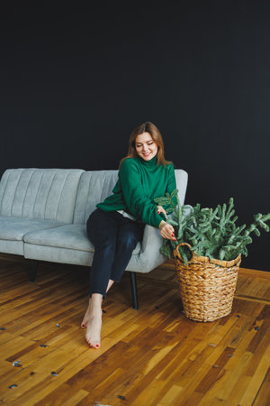 A happy woman in a green sweatshirt sits on a gray sofa, arranging vibrant greenery in a woven basket, creating a warm, tranquil home atmosphere for relaxationの写真素材