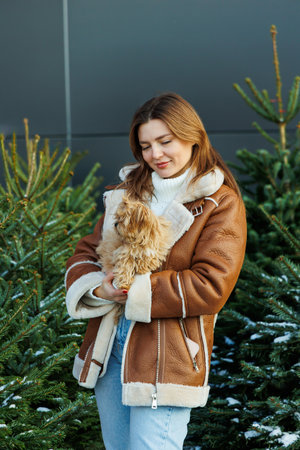 A cheerful young woman in trendy winter clothing holds a fluffy dog, surrounded by beautifully adorned evergreen trees, evoking warmth and happiness in this festive seasonの写真素材