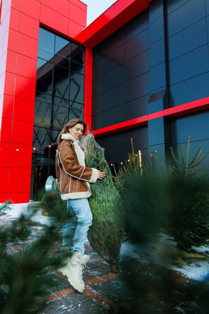 A cheerful woman in cozy winter attire embraces a beautifully decorated Christmas tree in front of a modern building, embodying the festive spirit of the seasonの写真素材