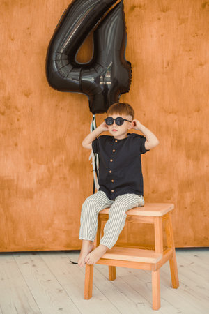 A cheerful young boy poses on a stylish chair, celebrating his 4th birthday in sunglasses and colorful pants, with a vibrant backdrop enhancing the festive atmosphereの写真素材
