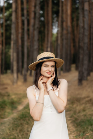 In a picturesque scene, a young woman in a flowing white dress and straw hat poses among lush green pine trees, embodying beauty and tranquility in this serene settingの写真素材